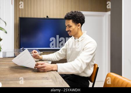 Un giovane professionista siede su una scrivania di legno, rivedendo i documenti con una penna in mano. L'ambiente moderno dell'ufficio, con schermo e arredi eleganti Foto Stock