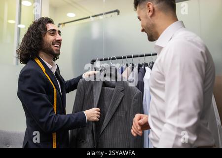 Un sarto aiuta un cliente a scegliere un abito in un negozio di abiti. Il sarto, con un metro a nastro, offre consigli esperti, presentando varie opzioni eleganti f Foto Stock