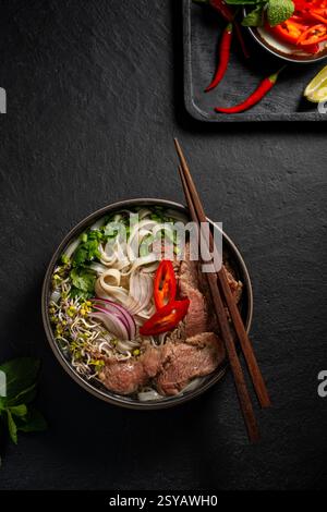 Vista dall'alto di una deliziosa ciotola di zuppa vietnamita di tagliatelle di manzo, con teneri fette di manzo, tagliatelle di riso piatte, erbe fresche come coriandolo e bas Foto Stock