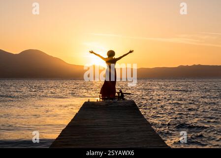 Una donna si erge su un molo di legno presso la spiaggia di Papikinou, l'isola di Milos, in Grecia, abbracciando il sole mentre tramonta, gettando un bagliore dorato sul mare tranquillo. Foto Stock