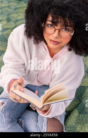Una giovane donna latina con gli occhiali ama leggere un libro seduto comodamente. Il suo stile casual e la sua attenzione creano un'atmosfera rilassata e intellettuale Foto Stock
