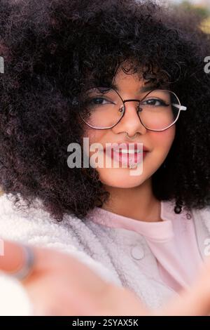 Una giovane donna latina con voluminosi capelli ricci, che indossa occhiali rotondi e un comodo maglione, sorride calorosamente. I suoi piercing facciali le conferiscono un tocco moderno Foto Stock