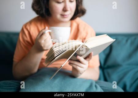 Vista corta di una donna che sorseggia il tè e legge un libro, comodamente seduto su un divano. Foto Stock
