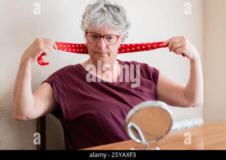 Una donna anziana con i capelli corti grigi e gli occhiali allacciano una sciarpa rossa a pois al collo. Si siede su un tavolo di legno con uno specchio rotondo, concentrato su Foto Stock