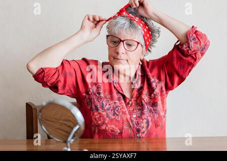 Una donna anziana con una camicia rossa vivace e floreale regola il suo velo rosso a pois mentre guarda in uno specchio su un tavolo di legno, creando un vivace ma allo stesso tempo Foto Stock