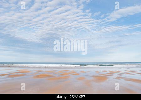 Una tranquilla spiaggia con un'ampia vista delle dolci onde sotto un cielo pieno di morbide nuvole, che mette in risalto la bellezza naturale di un undistur Foto Stock
