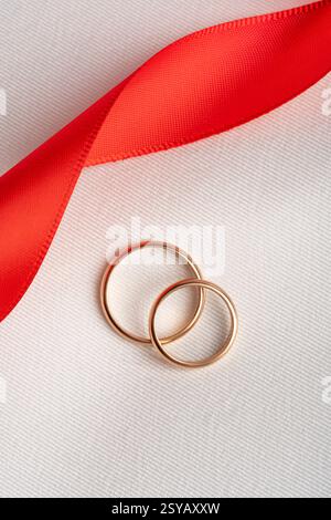 Vista dall'alto di due fedi nuziali dorate adagiate su un tessuto bianco testurizzato, intrecciato con un vivace nastro rosso, che simboleggia il romanticismo e l'amore eterno, id Foto Stock