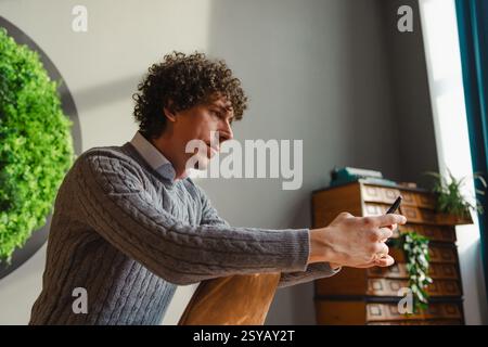 Un giovane con i capelli ricci usa uno smartphone, seduto accanto a una vivace parete verde. Questa scena mette in evidenza temi di ecologia e sviluppo sostenibile Foto Stock