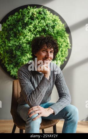 Un giovane uomo con i capelli ricci seduto con cura, incorniciato da una lussureggiante parete verde. Questa immagine mette in risalto i temi dell'ecologia e dello sviluppo sostenibile in Am Foto Stock