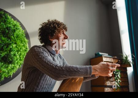 Un giovane con i capelli ricci siede in casa, sorridendo con un telefono in mano. Una parete verde circolare mette in risalto i temi dell'ecologia e dello sviluppo sostenibile Foto Stock