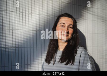 Una donna di mezza età sorride dolcemente mentre si trova in piedi contro una parete di piastrelle geometricamente modellata, con ombre diagonali che proiettano la luce del sole. Foto Stock