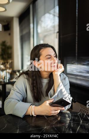 Una donna di mezza età siede pacificamente in un bar, guardando fuori mentre tiene in mano uno smartphone. La luce del sole mette in risalto la sua espressione premurosa Foto Stock