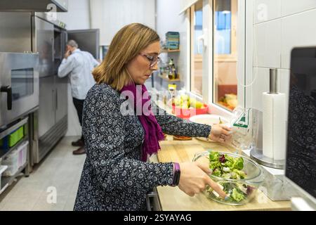 Una donna prepara un'insalata fresca in una cucina moderna, utilizzando una ciotola di vetro. Il vostro partner può essere visto in sottofondo, migliorando il vivace bancomat nazionale Foto Stock
