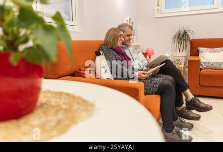 Una coppia con disabilità intellettive si diverte a rilassarsi insieme in una casa sorvegliata, leggendo su un comodo divano arancione. Una pianta rossa in vaso Foto Stock