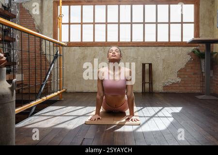 Una donna esegue yoga in una sala ben illuminata con luce naturale che scorre attraverso grandi finestre. L'ambiente rustico aggiunge un'atmosfera tranquilla Foto Stock