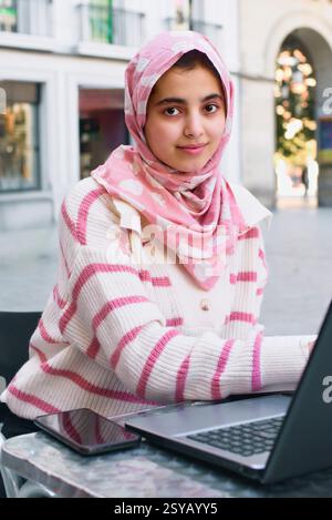 Una studentessa araba in un hijab rosa e maglione a righe utilizza un computer portatile in un bar all'aperto. Il suo smartphone si trova nelle vicinanze, incarnando una miscela di tradizione Foto Stock