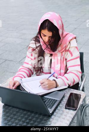Una studentessa araba che indossa un hijab rosa studia diligentemente a un tavolo all'aperto, circondato da un portatile e un notebook, illustrando la concentrazione e il determine Foto Stock