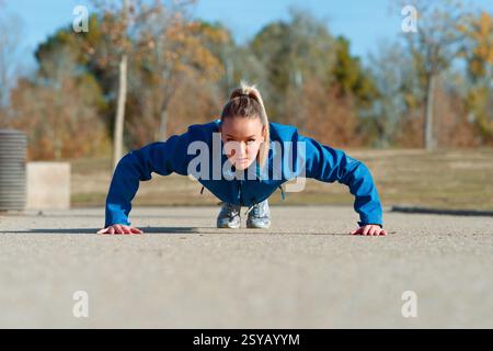 Una donna con una giacca blu esegue una spinta su una superficie in cemento all'esterno, concentrata e determinata. Lo sfondo mostra gli alberi autunnali sotto un bl chiaro Foto Stock