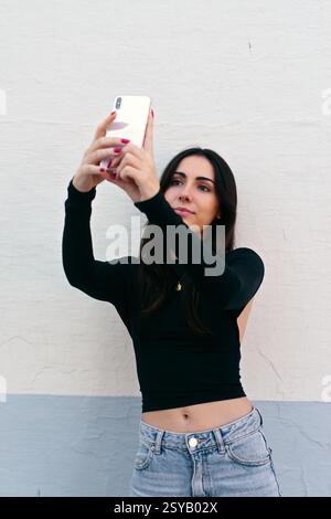 Una giovane donna con un top nero e jeans cattura un selfie con il suo smartphone. Si erge contro un muro bianco testurizzato, trasudando fiducia casuale in un Foto Stock