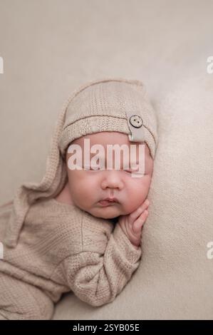 Un neonato sereno dorme tranquillamente mentre indossa un morbido abito in maglia beige, completato da un cappello coordinato con un grazioso bottone. La scena trasuda Foto Stock