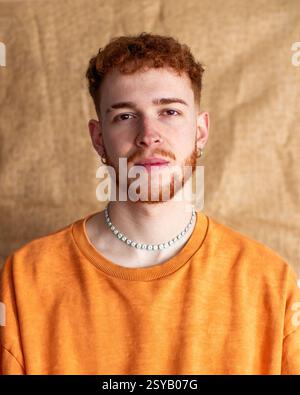 Un giovane con la barba rossa si posa con sicurezza in un maglione arancione e in una collana di perle. Lo sfondo semplice accentua il suo look casual ed elegante in t Foto Stock