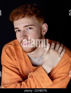 Ritratto di un giovane con i capelli rossi, con un maglione arancione e gioielli. Guarda penosamente la telecamera, mano sulla spalla, contro un dorso scuro Foto Stock