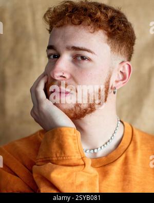 Un ritratto ravvicinato di un giovane uomo alla moda con i capelli ricci rossi, che indossa una collana di perle e orecchini. Guarda con attenzione, con la mano sul suo che Foto Stock