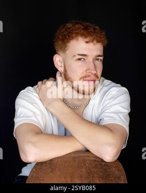 Ritratto di un giovane uomo con i capelli rossi e un'espressione premurosa, seduto su uno sfondo nero. Indossa una camicia bianca e una collana d'argento, annuncio Foto Stock