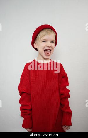 Un bambino allegro con un comodo maglione rosso e un berretto abbinato ride gioiosamente su uno sfondo semplice. L'immagine cattura un momento di felicità spensierata Foto Stock