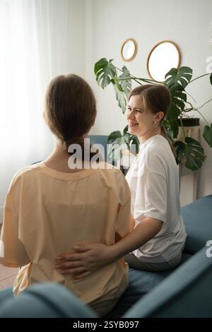 Una madre e sua figlia condividono un momento caldo sul divano, circondati da piante. Le loro espressioni rilassate e l'atmosfera accogliente mettono in risalto l'imp Foto Stock