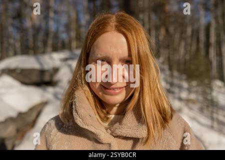 Una donna sorridente di capelli rossi che guarda la fotocamera in un'accogliente giacca si trova in una foresta innevata, crogiolandosi alla luce del sole di una fresca giornata invernale. Il clima è ev Foto Stock