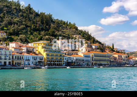 Vista pittoresca delle colorate case sul lungomare di Gythio e degli affascinanti edifici lungo la costa, con lussureggianti colline verdi sullo sfondo sotto una luce luminosa Foto Stock