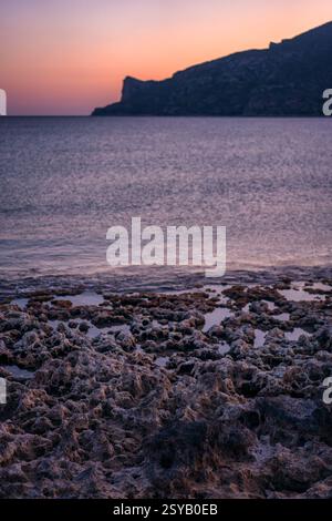 Un tranquillo paesaggio marino greco al tramonto, caratterizzato da rocce aspre sulla spiaggia e calme acque color viola, con una montagna sagomata in lontananza un Foto Stock