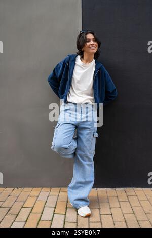 Una persona si appoggia a una parete bicolore, indossando un outfit casual con felpa con cappuccio blu, T-shirt bianca, jeans azzurri e sneakers bianche. La scena exu Foto Stock