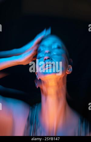 Un suggestivo ritratto di una donna calva, catturato con una vivace luce blu e arancione. La sua posa espressiva trasmette un senso di movimento ed emozione, enhanc Foto Stock