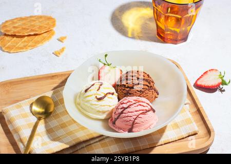Delizioso trio di gelati su un vassoio di legno con cucchiaio dorato e biscotti a base di waffle, catturato da un angolo leggermente alto con luce naturale brillante Foto Stock