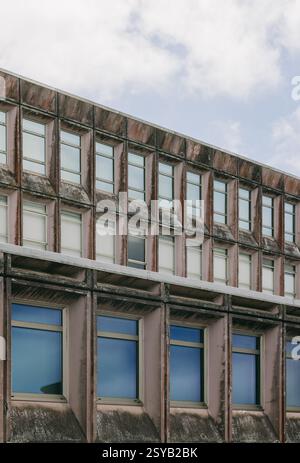 Foto ravvicinata di una facciata di un edificio dall'architettura brutalista, caratterizzata da file di finestre contro un cielo nuvoloso. Struttura in cemento invecchiato displ Foto Stock