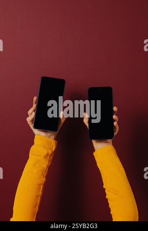 Vista laterale di una persona irriconoscibile in abiti casual che tengono in mano smartphone con schermo nero mentre si sta in piedi su sfondo rosso scuro Foto Stock