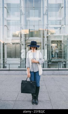 Una donna alla moda si erge di fronte a un moderno edificio in vetro con un cappello a teglia larga, jeans blu e un gilet bianco. Ha una grossa mano nera Foto Stock