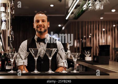 Un barista professionista si erge con fiducia dietro un bar elegante adornato con bicchieri da martini, trasudando competenza e ospitalità in un cocktail moderno Foto Stock