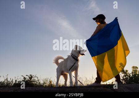 donna che tiene una grande bandiera ucraina dietro di lei e un cane accanto a lei contro il cielo del tramonto. cammina con l'amato animale domestico nel giorno dell'indipendenza ucraina. orgoglio di essere u Foto Stock