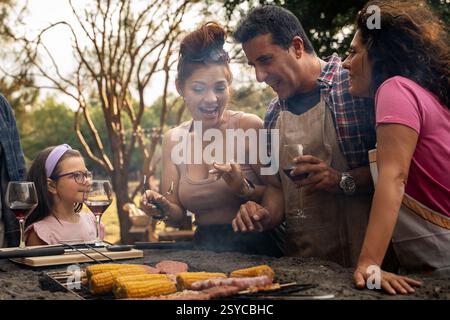 Una famiglia allegra e amici che si godono un barbecue all'aperto, condividono cibo e risate in un ambiente rustico con mais alla griglia e vino. Foto Stock
