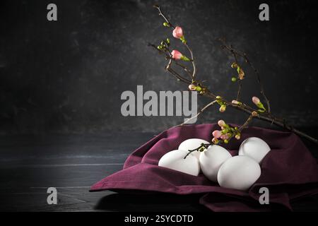 Uova bianche su un tovagliolo viola e un ramoscello di cotogna con gemme rosa su sfondo grigio scuro, decorazione di Pasqua chiara al buio, spazio copia, sele Foto Stock