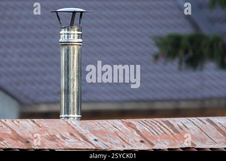 Un tubo metallico del camino con un tappo si erge su un tetto rosso sotto uno sfondo del tetto fuori fuoco Foto Stock