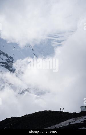 Tiro verticale di due uomini che lavorano sotto enormi montagne innevate avvolte da nuvole, Pamir, Kirghizistan Foto Stock