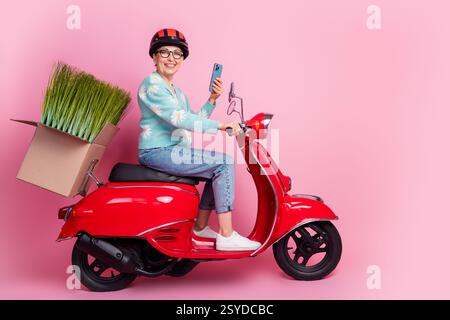 Donna anziana gioiosa seduta su uno scooter rosso con una scatola decorativa su sfondo rosa Foto Stock