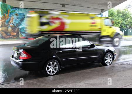 Berlina Mercedes-Benz nera parcheggiata sotto un cavalcavia con un camion giallo in movimento e arte murale sullo sfondo in una giornata di pioggia. Foto Stock