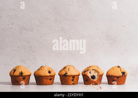 Una fila di muffin dorati ai mirtilli dall'estetica rustica e minimalista, con un boccone che rivela un interno soffice e pieno di frutti di bosco. Foto Stock