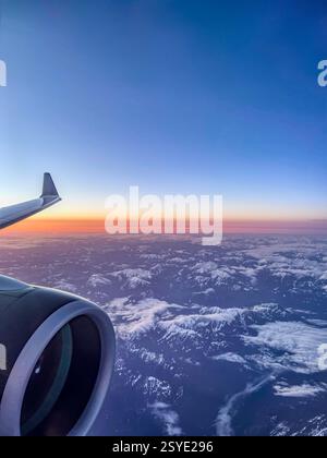 Vista aerea dalla finestra di un aereo che mostra l'ala e il motore su una catena montuosa innevata all'alba. Il cielo vibrante passa dal blu intenso Foto Stock