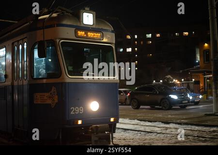 Un tram blu è illuminato di notte nel centro della città, con la sua destinazione visualizzata. Le auto parcheggiate fiancheggiano la strada innevata, e gli edifici sono visibili io Foto Stock